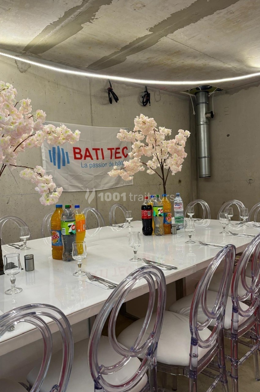 Table dressée avec des chaises transparentes, boissons variées et décorations florales dans une salle au mur brut.