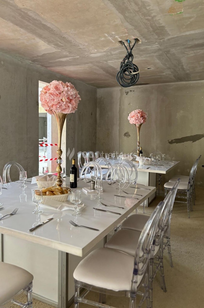 Salle de réception en travaux avec tables dressées, chaises transparentes et centres de table ornés de fleurs roses.