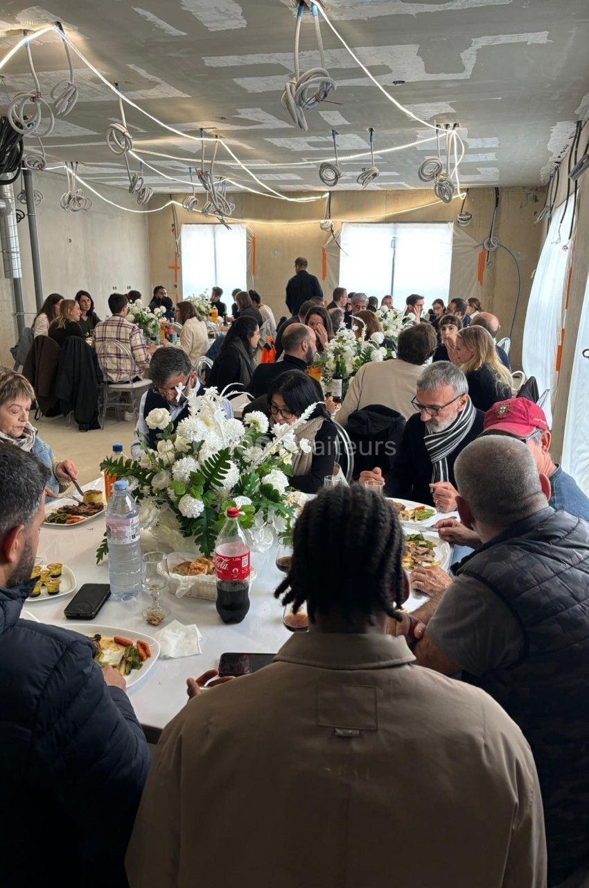 Des convives assis autour de grandes tables décorées de fleurs blanches, partageant un repas dans une salle en travaux.