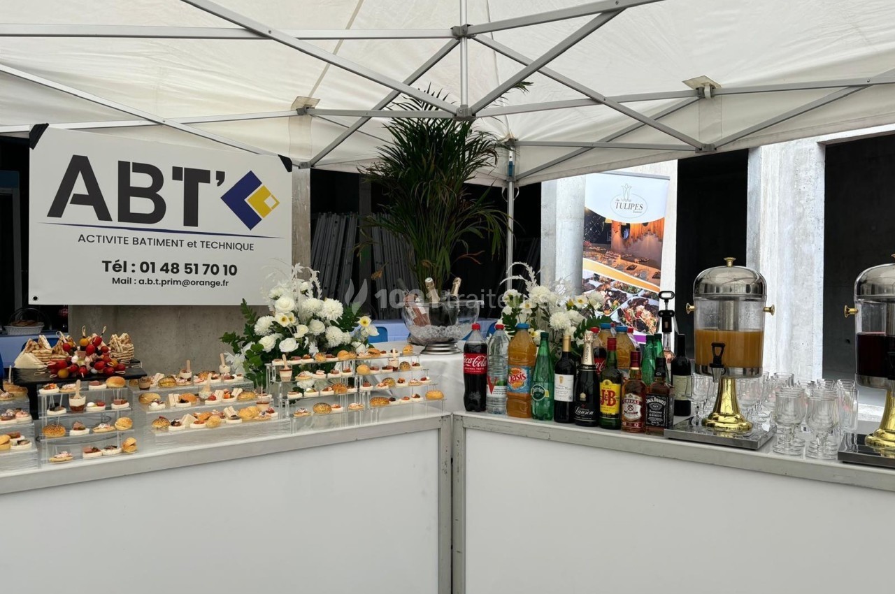 Buffet sous une tente avec boissons, amuse-bouches et décorations florales, devant un panneau d'entreprise ABT.