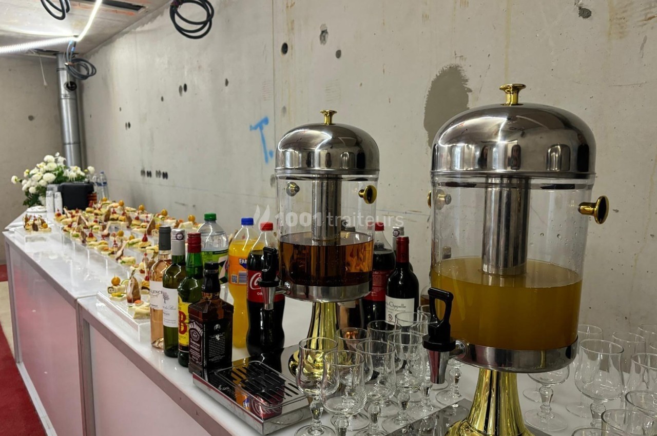 Buffet avec boissons variées, distributeurs de jus, verres et collations disposés sur une table dans un espace brut.