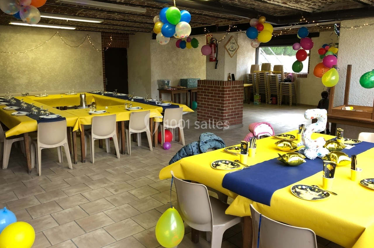 Salle décorée pour une fête, avec tables dressées en jaune et bleu, ballons colorés et guirlandes lumineuses.