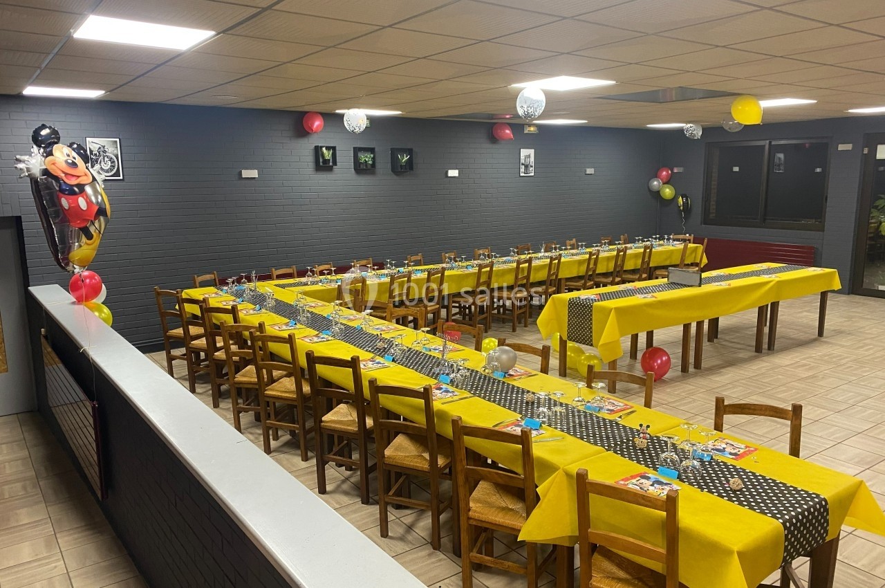 Salle décorée pour une fête, avec des tables alignées, nappes jaunes, ballons et un thème enfantin.