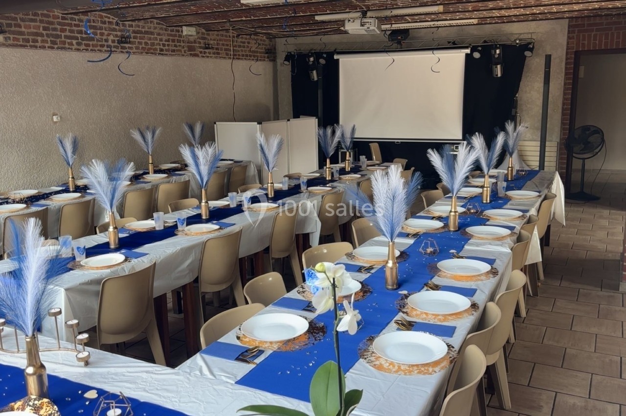Salle décorée pour un événement, avec de longues tables dressées, nappes bleues et blanches, et centres de table ornés de…
