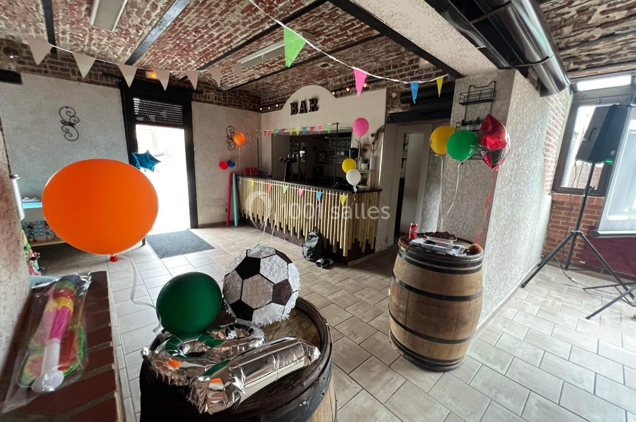 Salle décorée pour une fête avec ballons colorés, guirlandes et un bar en bois entouré de tonneaux.