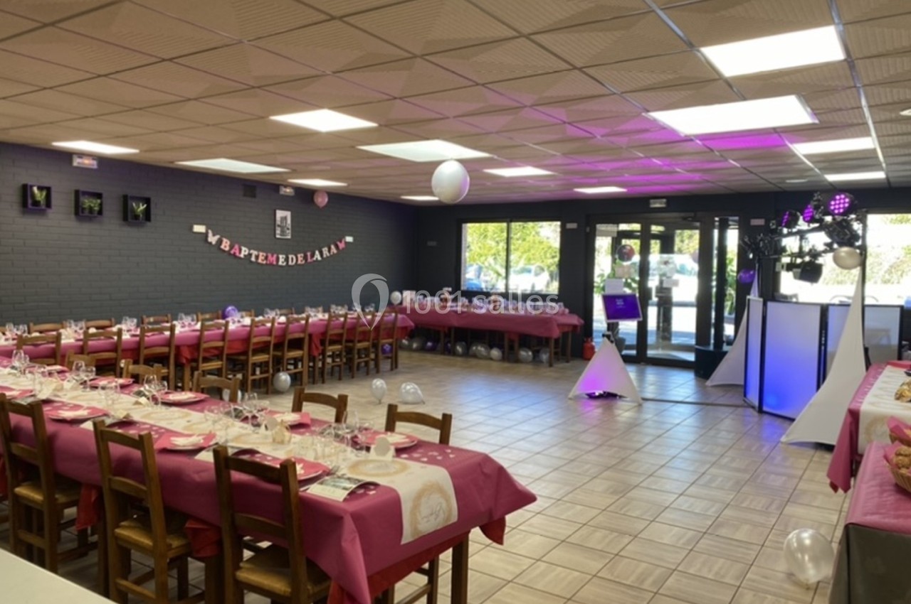 Salle décorée pour un baptême avec tables dressées en rose, guirlande murale et espace DJ éclairé.