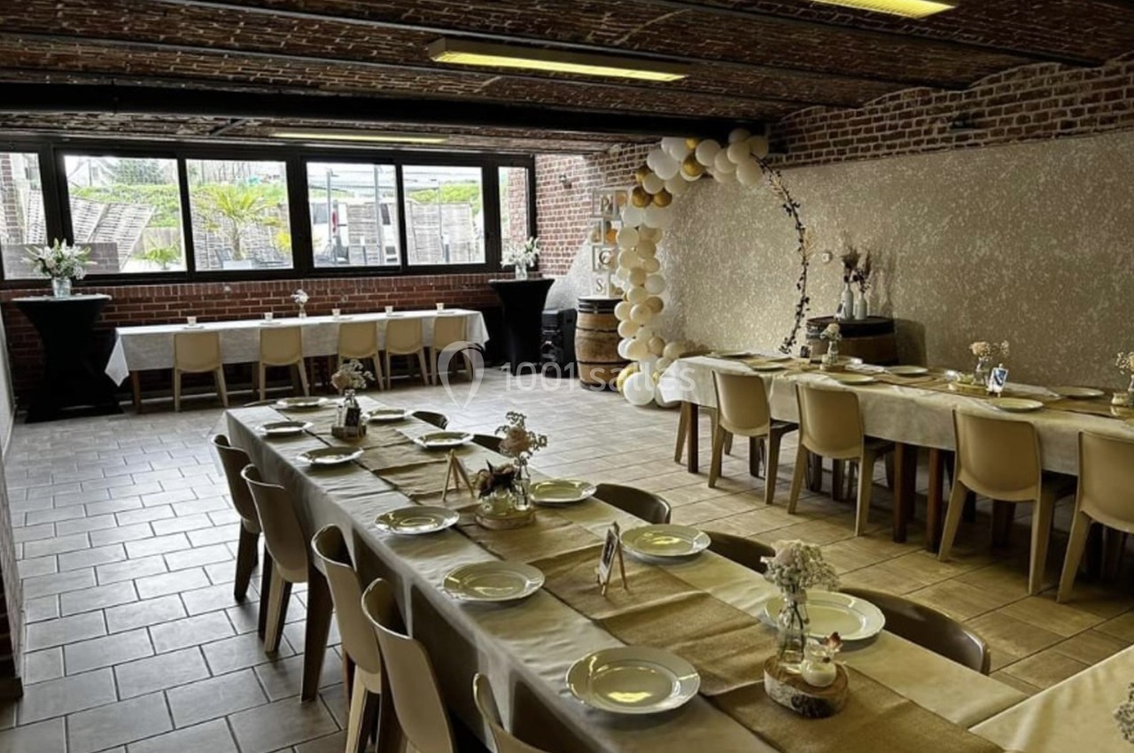 Salle décorée pour un événement, avec tables dressées, chaises beiges et arche de ballons près d'un mur en briques.