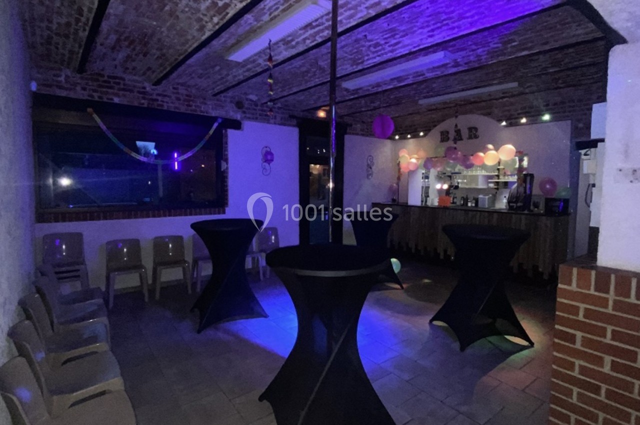 Salle avec tables hautes noires, chaises alignées, éclairage tamisé et bar décoré de guirlandes lumineuses.