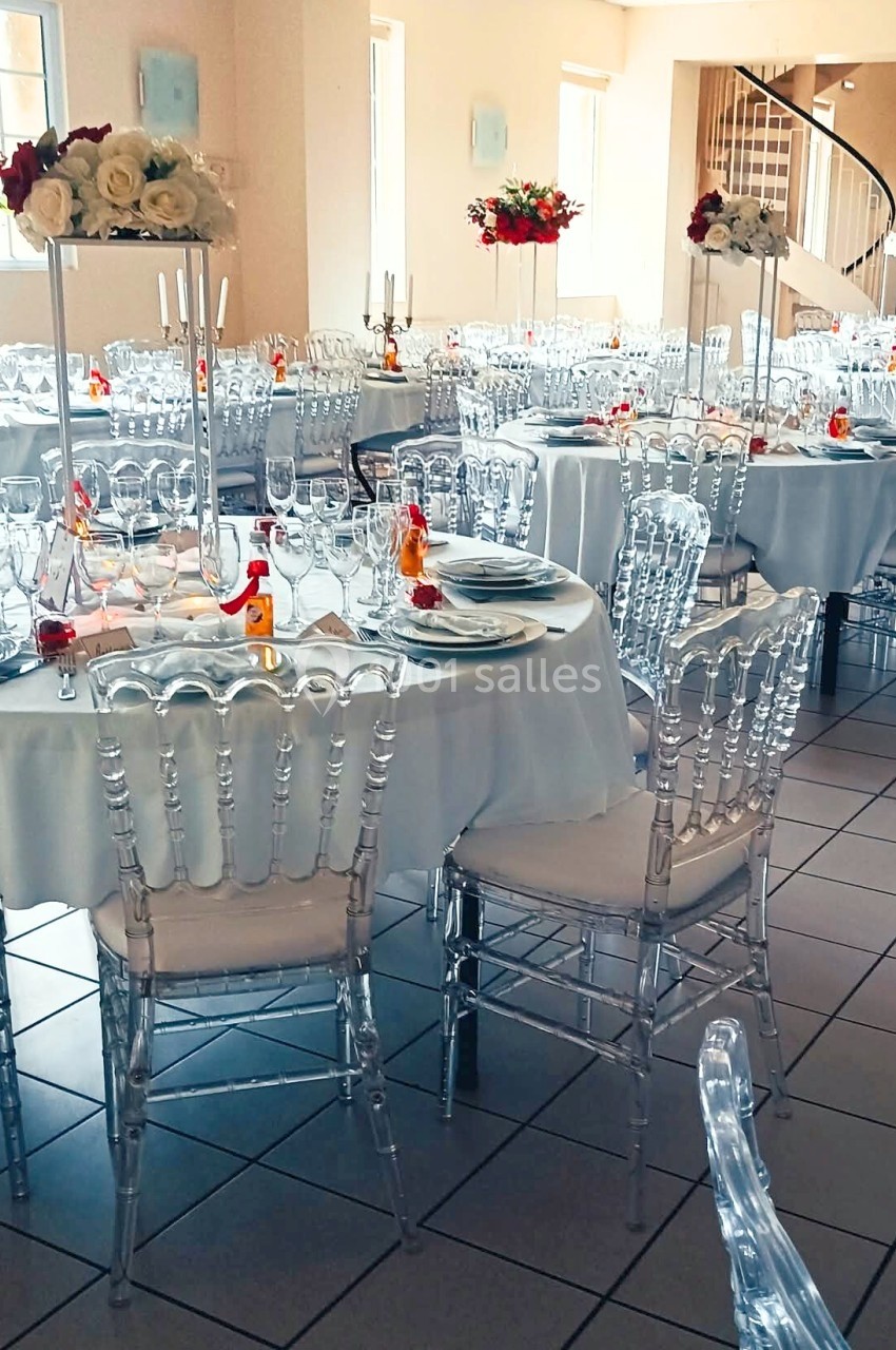 Salle de réception décorée avec des tables rondes, nappes blanches, chaises transparentes et centres de table fleuris.
