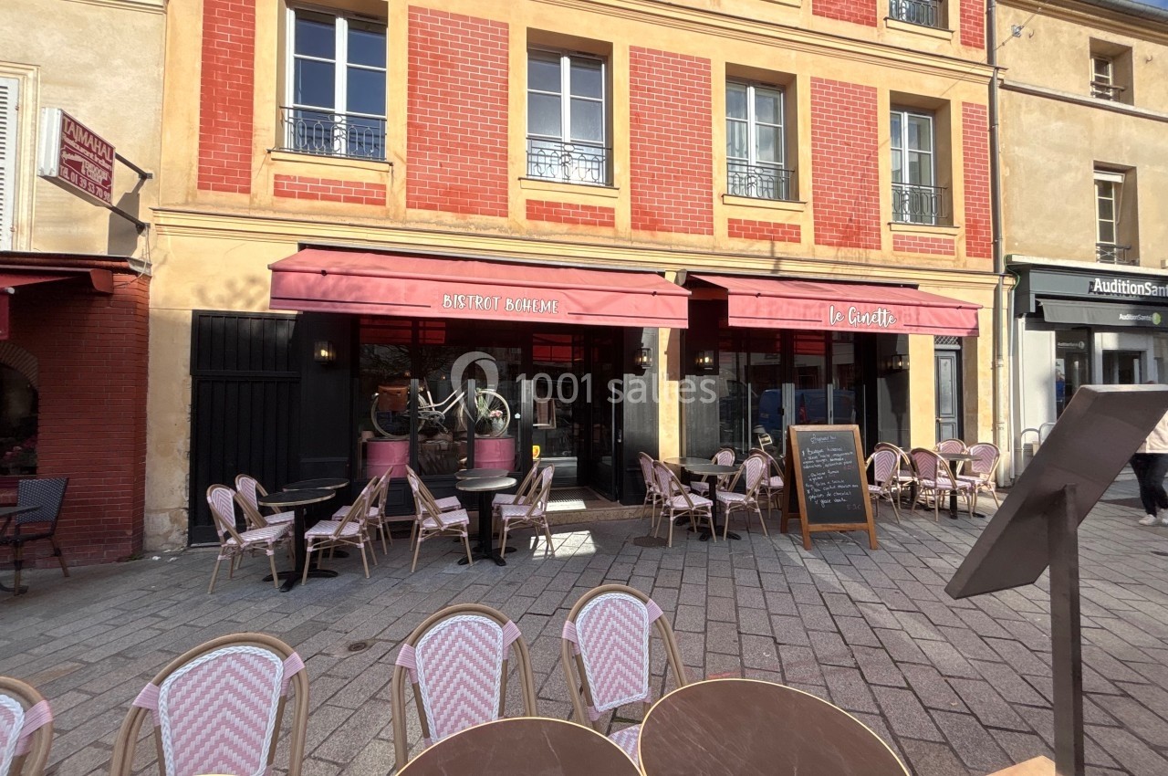 Façade d'un bistrot avec terrasse extérieure, chaises en osier et ardoise de menu sur une place pavée.