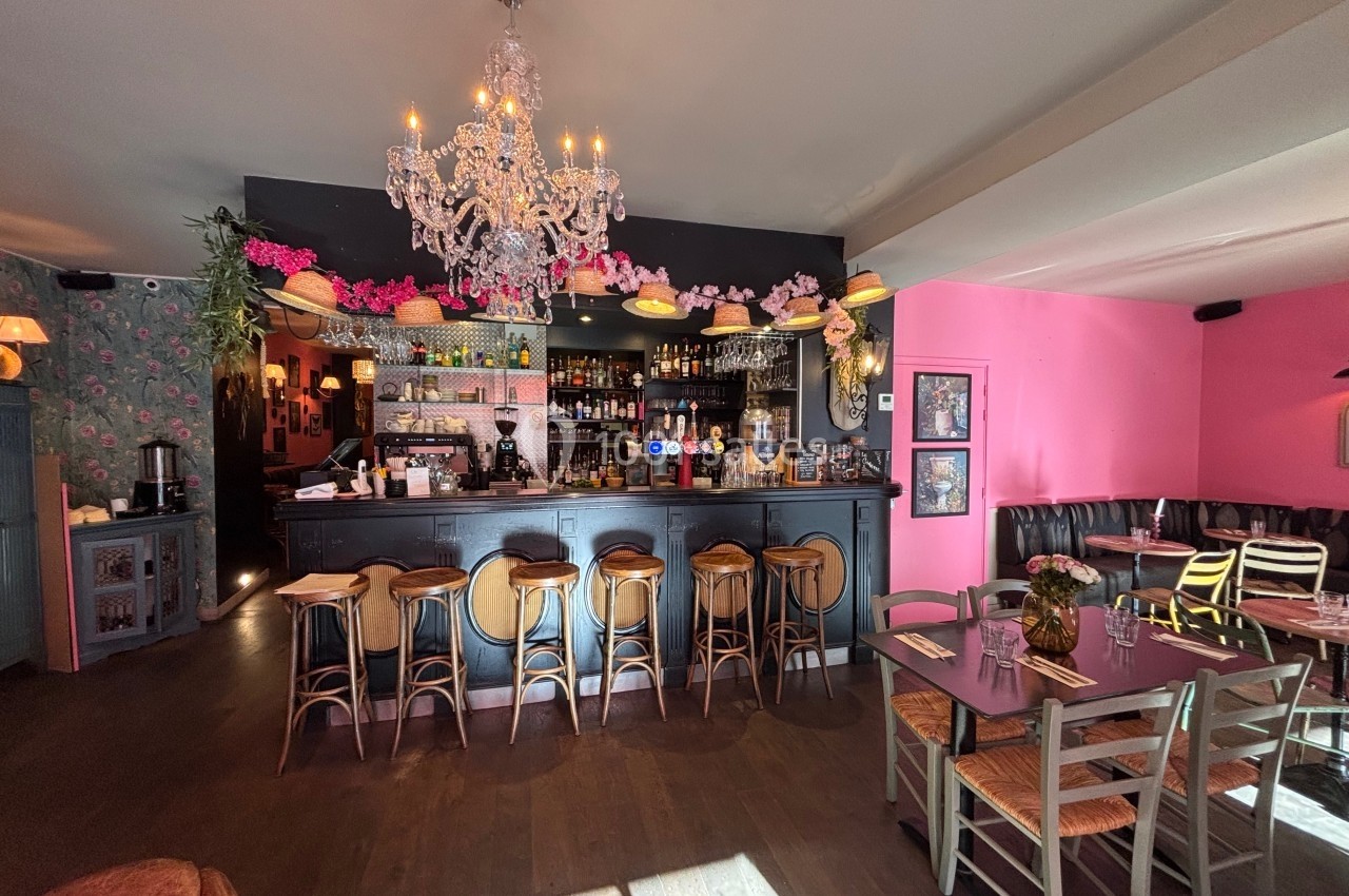 Intérieur d'un bar-restaurant avec comptoir noir, chaises en bois, lustre, murs roses et tables dressées.