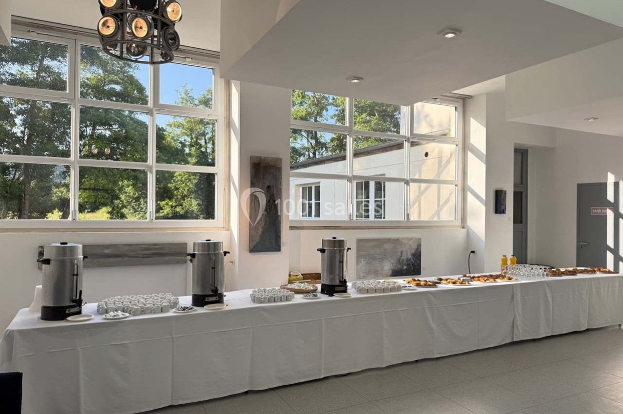 Table dressée avec boissons chaudes, jus et viennoiseries dans une salle lumineuse avec de grandes fenêtres.