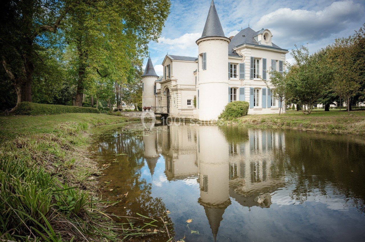 Château blanc avec tours, entouré d'arbres, reflété dans un étang calme sous un ciel partiellement nuageux.