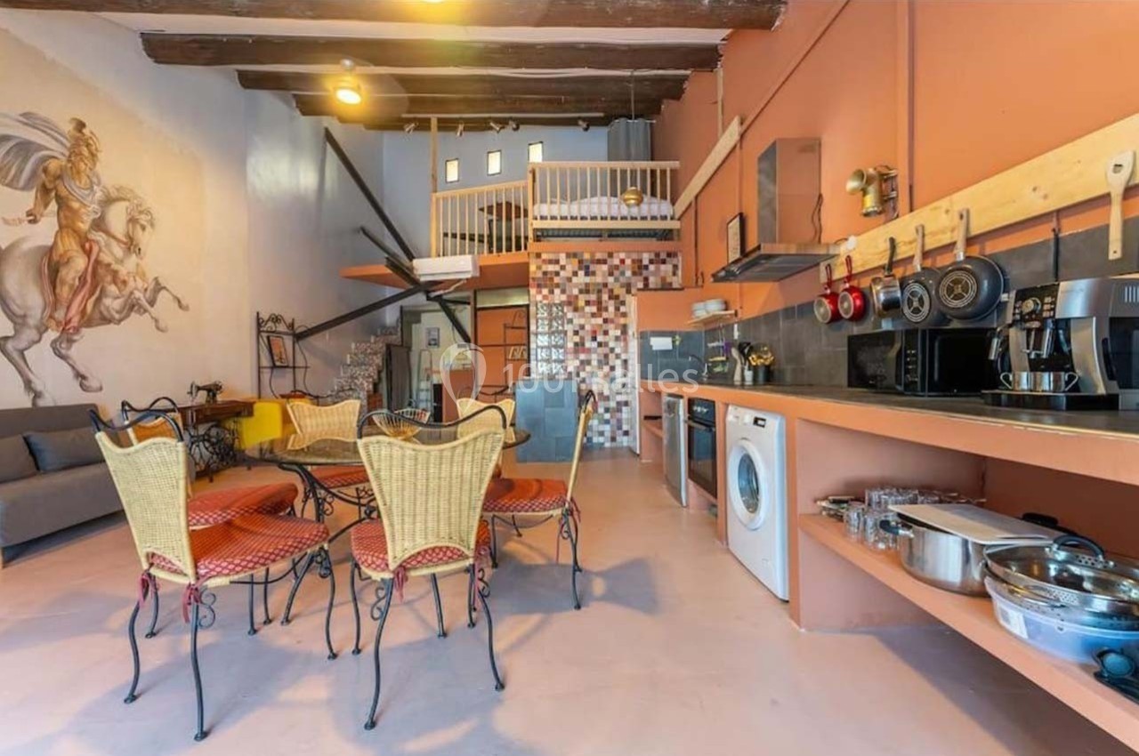 Espace de vie lumineux avec cuisine équipée, table à manger, canapé, mezzanine et décoration murale artistique.