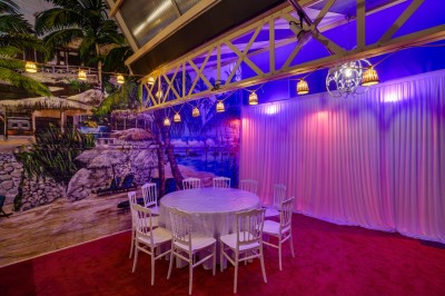 Salle décorée avec des tables rondes et chaises blanches, mur peint représentant une plage avec cabane et palmiers.