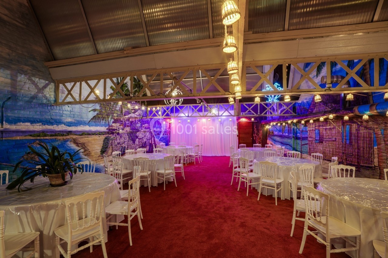 Salle décorée avec des tables rondes, chaises blanches, murs peints de paysages tropicaux et éclairage tamisé.