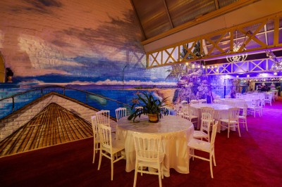 Salle décorée avec des tables rondes et chaises blanches, mur peint représentant une plage avec cabane et palmiers.
