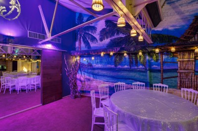 Salle décorée avec des tables rondes et chaises blanches, mur peint représentant une plage avec cabane et palmiers.