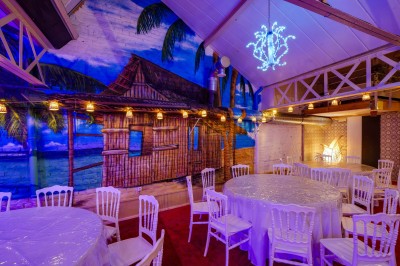 Salle décorée avec des tables rondes et chaises blanches, mur peint représentant une plage avec cabane et palmiers.