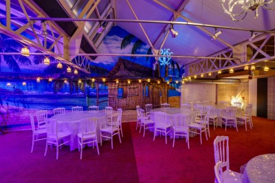 Salle décorée avec des tables rondes et chaises blanches, mur peint représentant une plage avec cabane et palmiers.