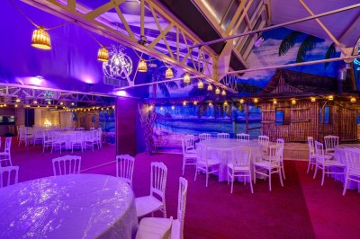 Salle décorée avec des tables rondes et chaises blanches, mur peint représentant une plage avec cabane et palmiers.