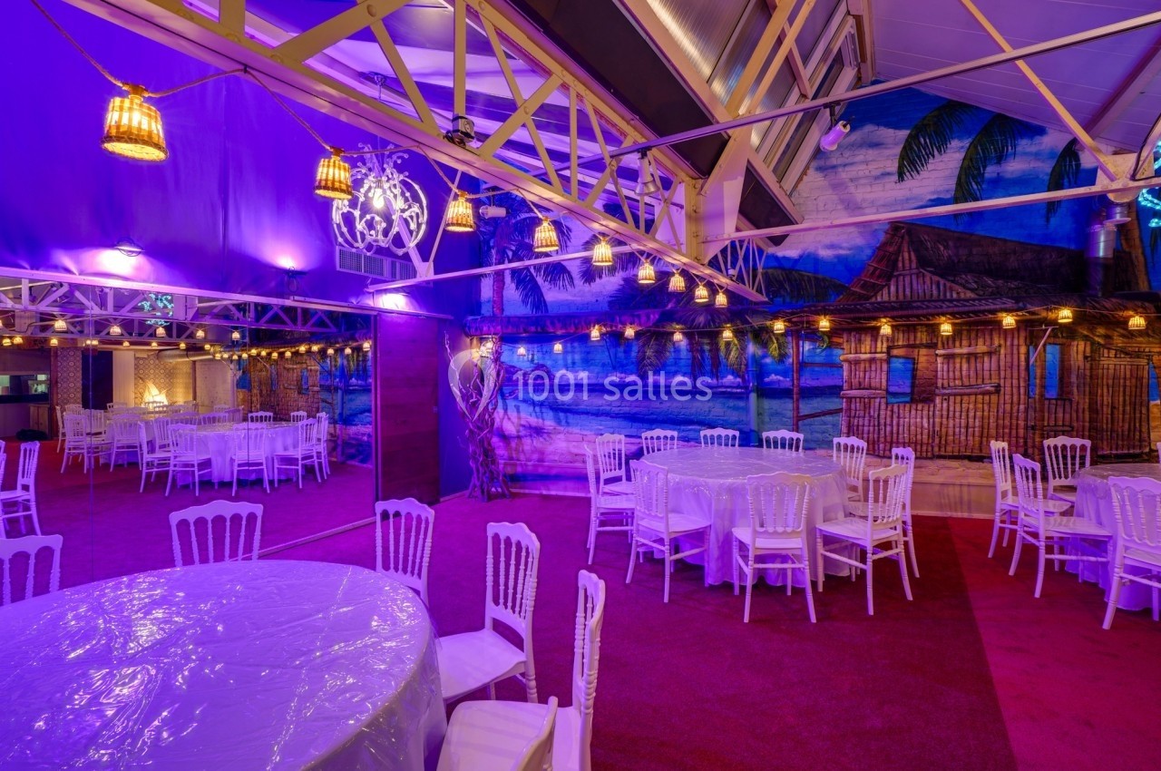 Salle décorée avec des tables rondes et chaises blanches, éclairage violet et fresque murale tropicale en arrière-plan.