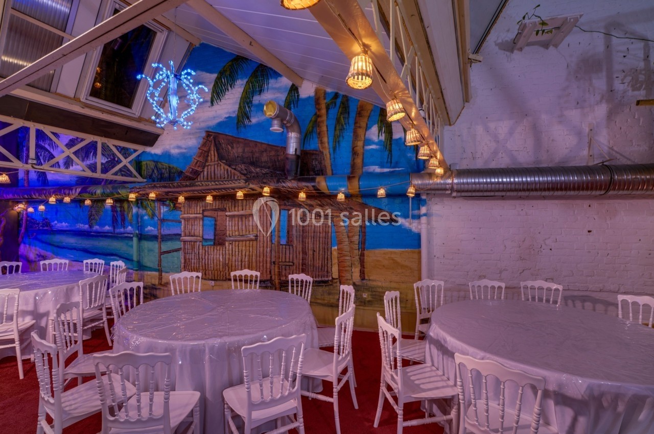 Salle décorée avec des tables rondes et chaises blanches, mur peint représentant une plage avec cabane et palmiers.