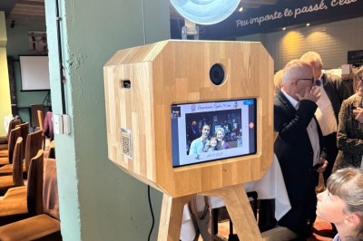 Borne photo en bois sur trépied, équipée d'un écran et d'un objectif, installée dans un jardin avec des coquelicots au…