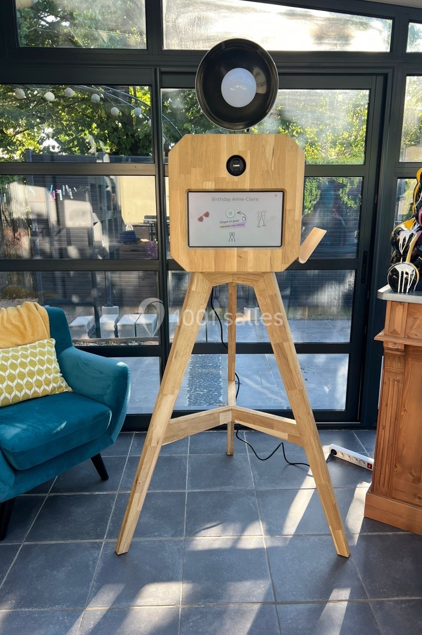 Borne photo en bois sur trépied avec écran tactile, installée dans une pièce lumineuse avec une baie vitrée.