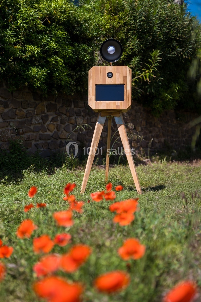 Borne photo en bois sur trépied, équipée d'un écran et d'un objectif, installée dans un jardin avec des coquelicots au…