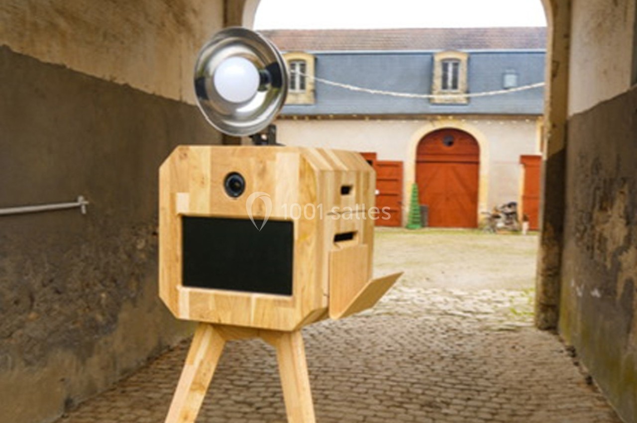 Boîte photo en bois sur trépied avec flash, placée dans une cour pavée devant des bâtiments anciens.