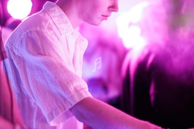 Un DJ mixe dans une ambiance de boîte de nuit avec des faisceaux lumineux et une foule floue en arrière-plan.
