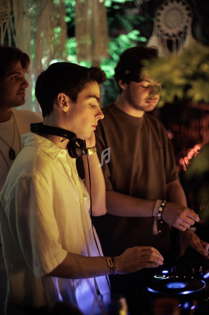 Trois DJ mixent de la musique en soirée, entourés de lumières tamisées et de décorations suspendues.