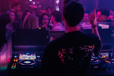 Un DJ mixe dans une ambiance de boîte de nuit avec des faisceaux lumineux et une foule floue en arrière-plan.