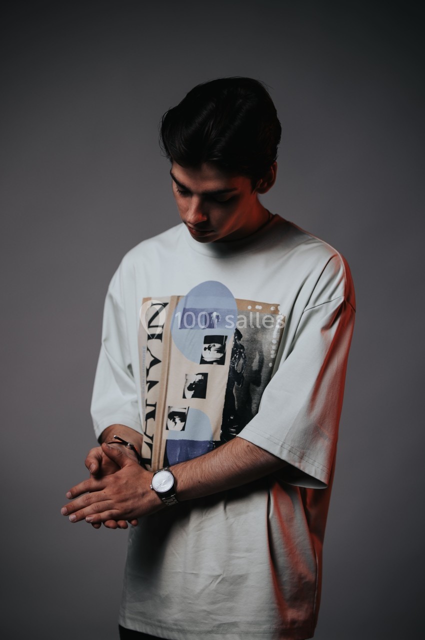 Un homme debout, regard baissé, portant un t-shirt imprimé et une montre, dans un éclairage tamisé.