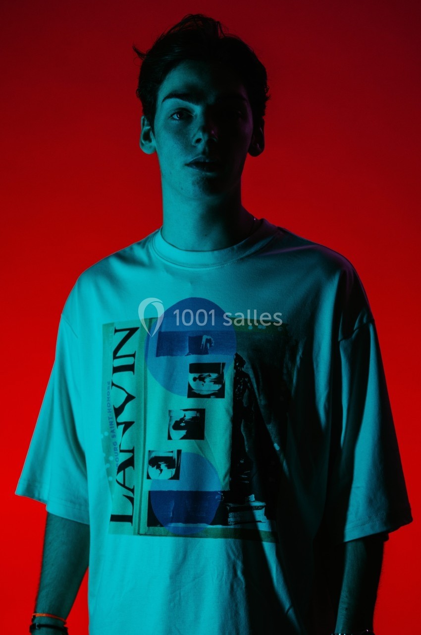 Un homme portant un t-shirt imprimé, éclairé par une lumière rouge et bleue dans un environnement sombre.
