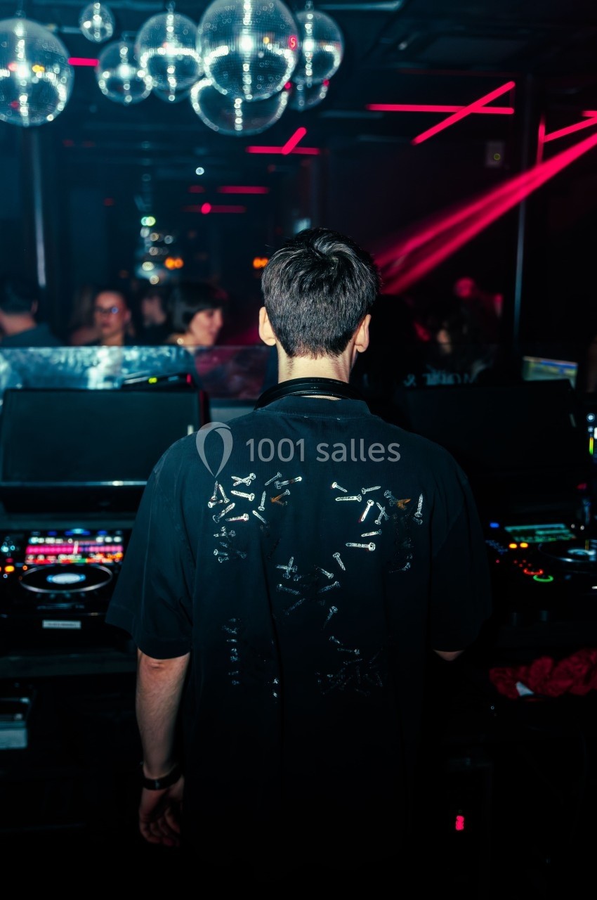 Un DJ de dos mixe dans une boîte de nuit sombre, éclairée par des lumières rouges et des boules à facettes.