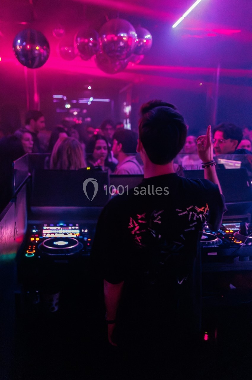 Un DJ mixe devant une foule dans une boîte de nuit éclairée par des lumières roses et des boules à facettes.