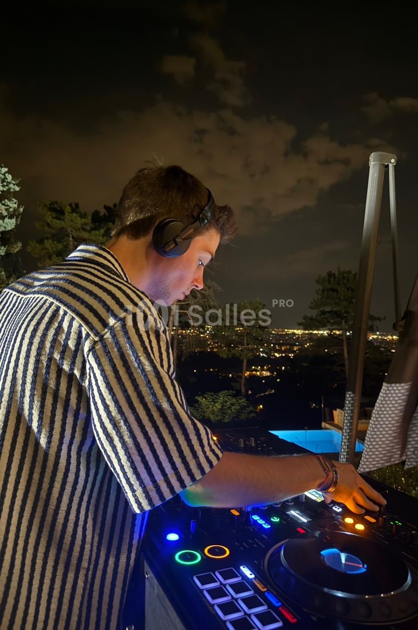 Un DJ mixe de la musique en extérieur la nuit, avec une vue sur une ville illuminée en arrière-plan.