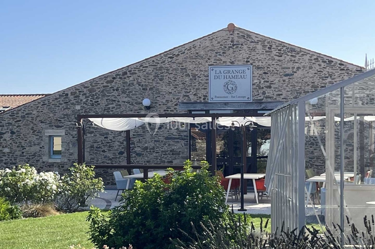Façade en pierre d'un restaurant avec terrasse couverte, entourée de verdure et de fleurs sous un ciel bleu.