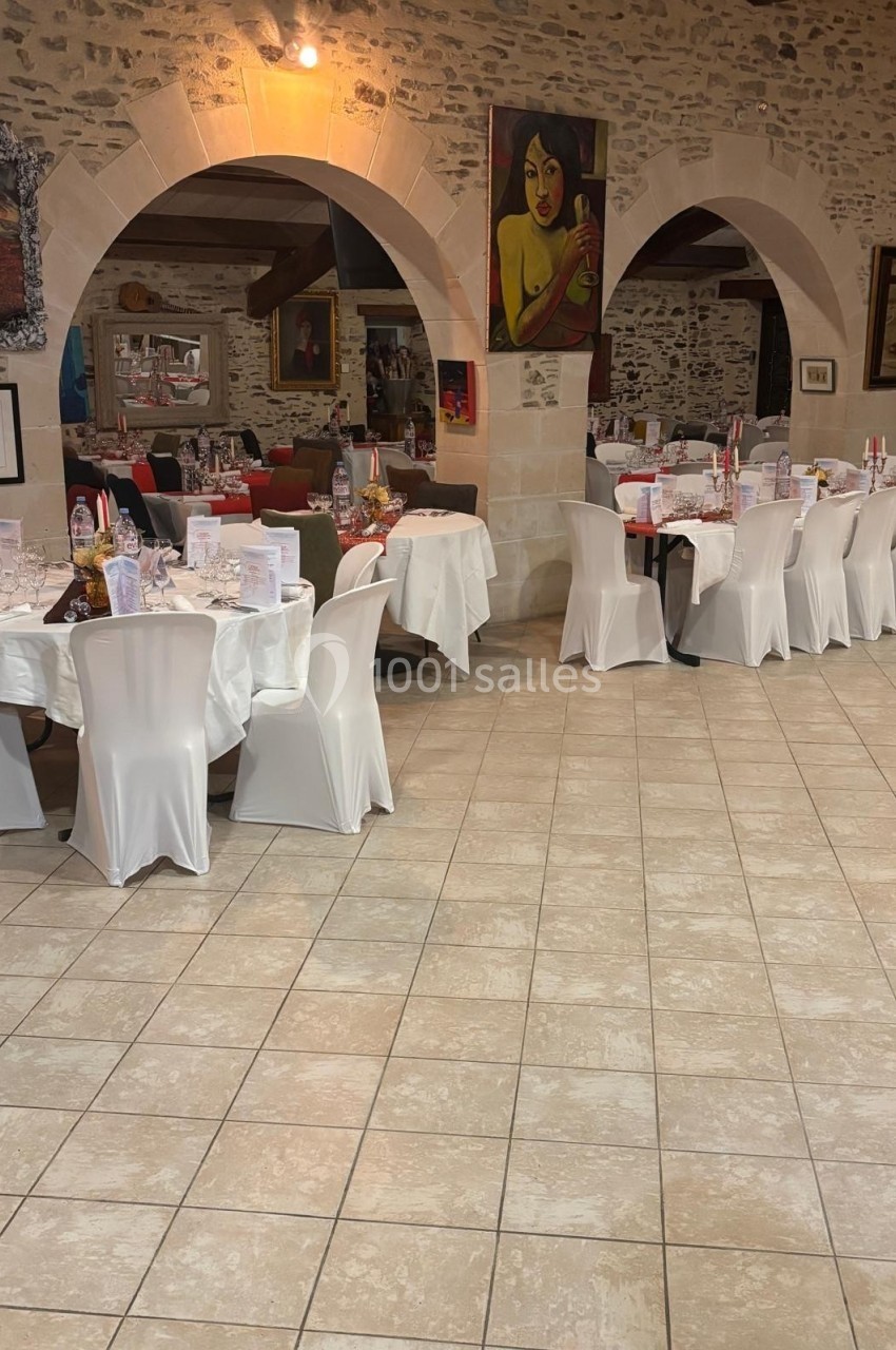 Salle de réception avec tables rondes et rectangulaires dressées, chaises blanches et murs en pierre apparente.
