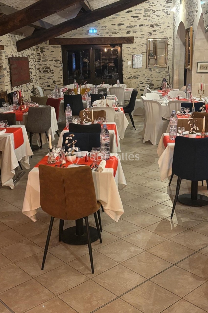 Salle de restaurant avec tables dressées, nappes blanches et rouges, chaises variées, murs en pierre et poutres apparentes.