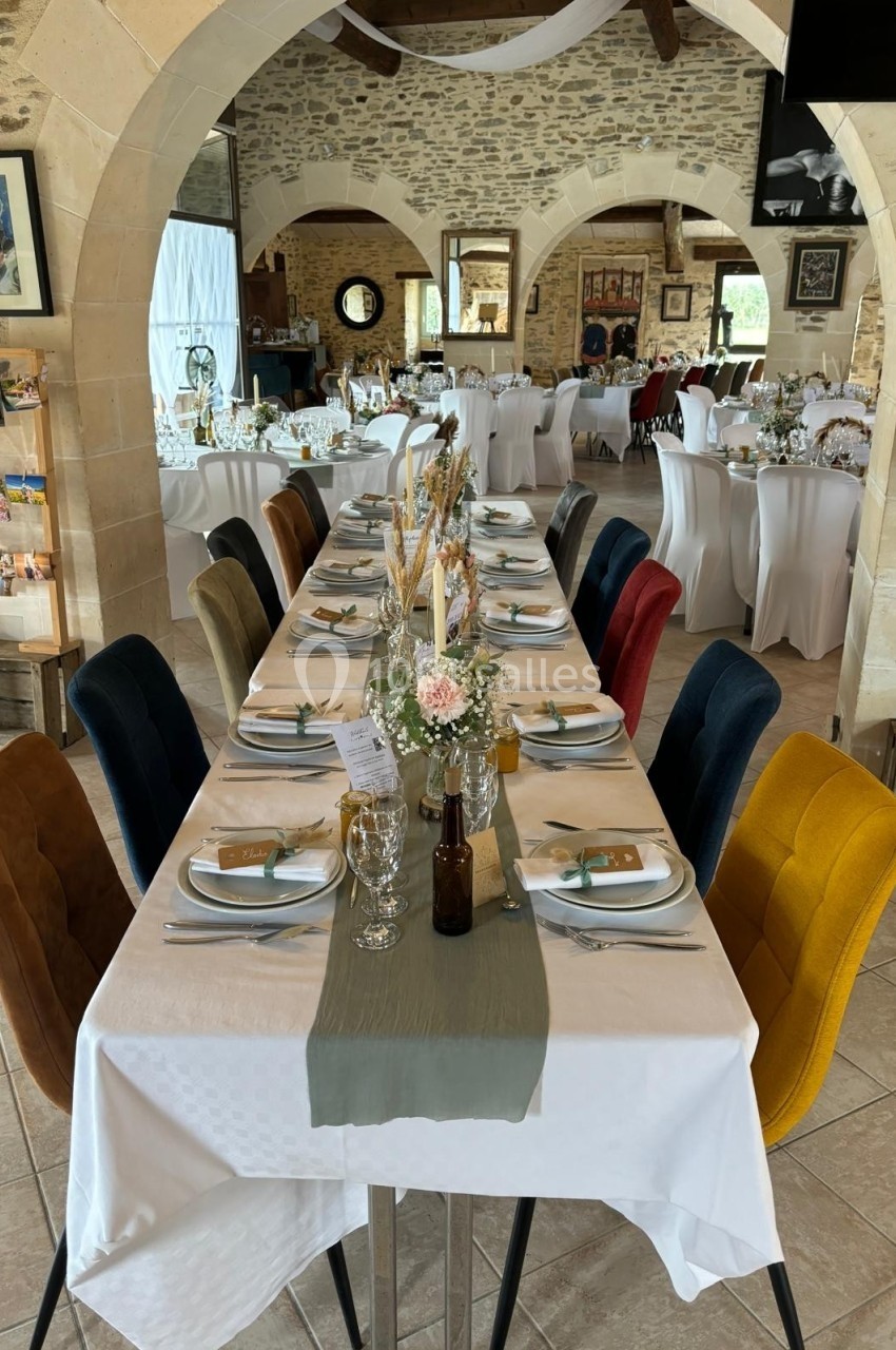 Salle de réception décorée avec des tables dressées, chaises colorées et éléments floraux, dans un cadre en pierre apparente.
