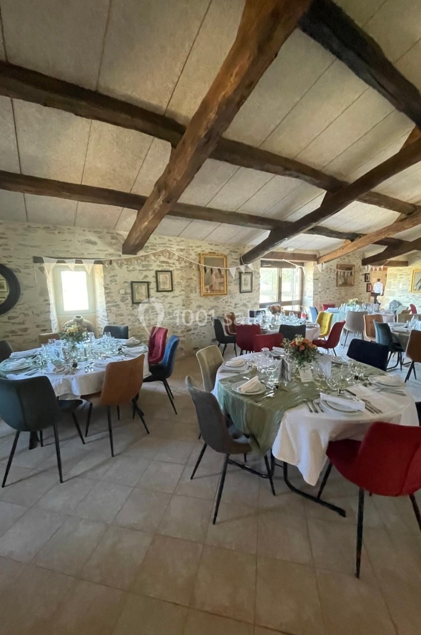 Salle de réception lumineuse avec tables rondes dressées, chaises colorées et poutres apparentes au plafond.
