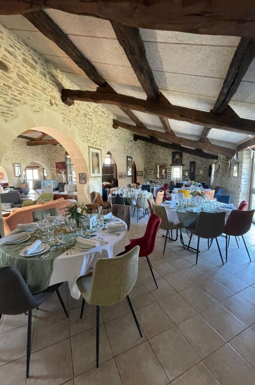 Salle en pierre avec poutres apparentes, tables dressées pour un repas, chaises colorées et lumière naturelle.