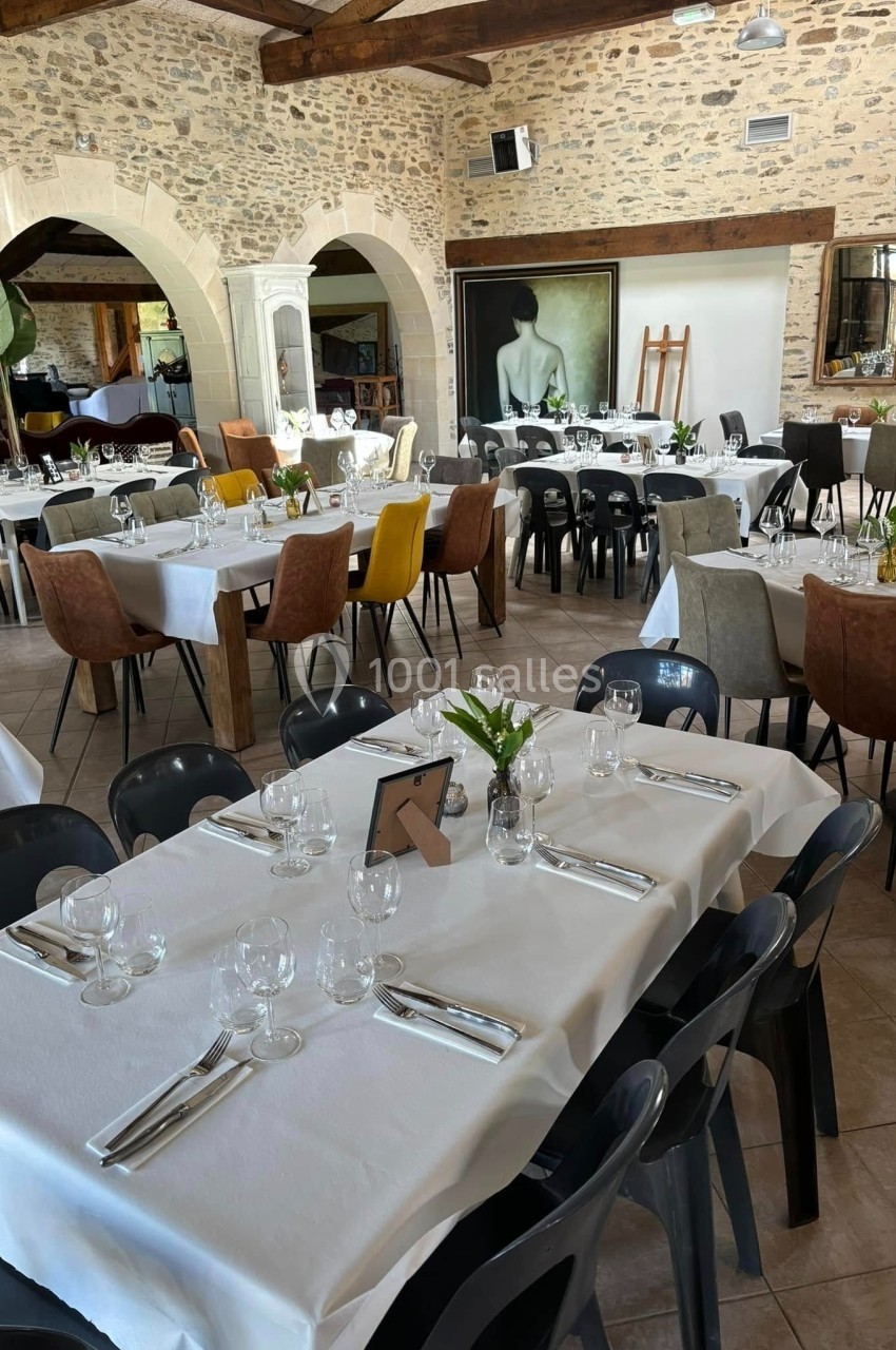 Salle de restaurant lumineuse avec tables dressées, nappes blanches, chaises variées et murs en pierre apparente.