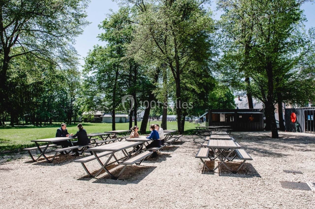Tables de pique-nique en bois disposées sous des arbres dans un espace extérieur ensoleillé avec quelques personnes assises.