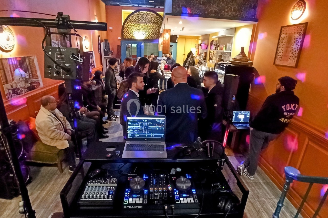 Vue d'une soirée dans un bar avec des invités debout, un DJ et des équipements de mixage au premier plan.