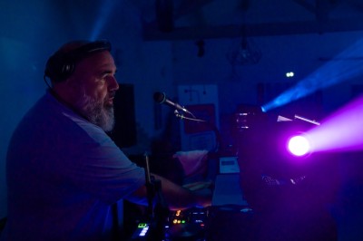 Miniature DJ Ventabren (Bouches-du-Rhône) - Provence Event #8 Un DJ en chemise rouge se tient derrière une table de mixage avec des lumières colorées en arrière-plan.