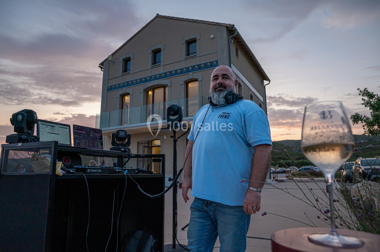 Un homme souriant devant une table de mixage en extérieur, avec une maison et un coucher de soleil en arrière-plan.